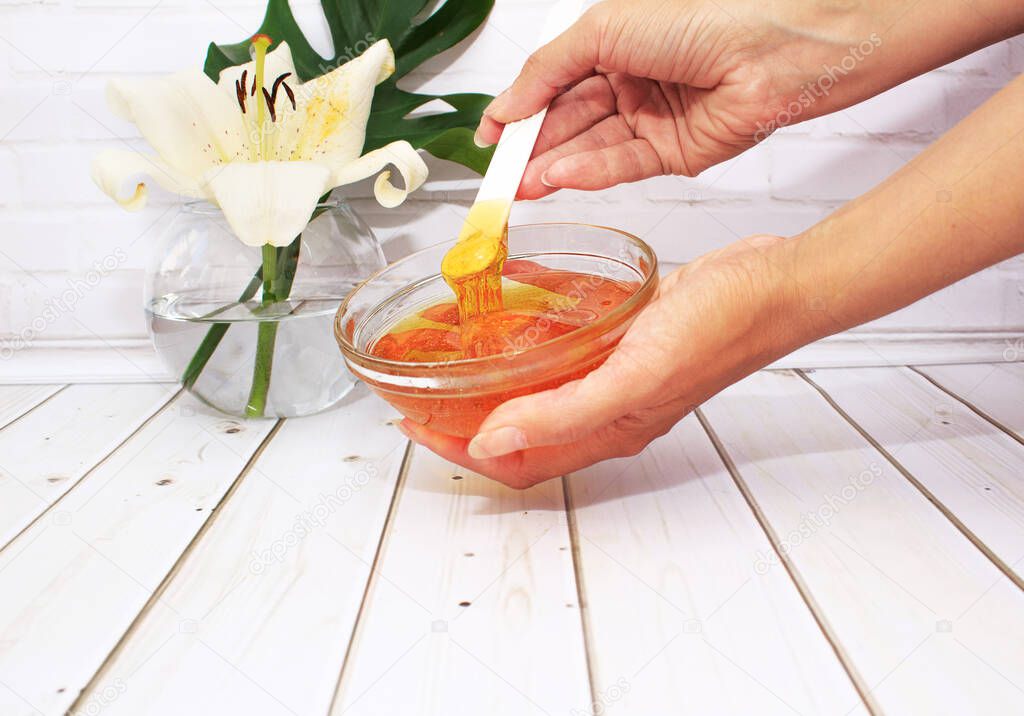 Femal hands hold liquid sugar paste for hair removal in a bowl with lily flower and dracaena leaf in glass vase. The concept of body care, beauty treatments.