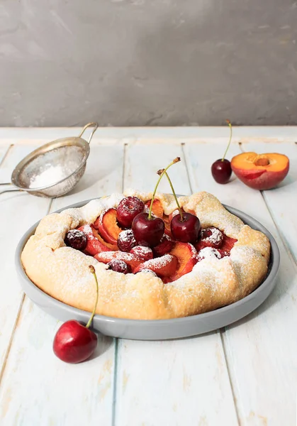 Homemade summer cake, galette with cherries and peaches with powdered sugar on a gray plate on a wooden background. Close up