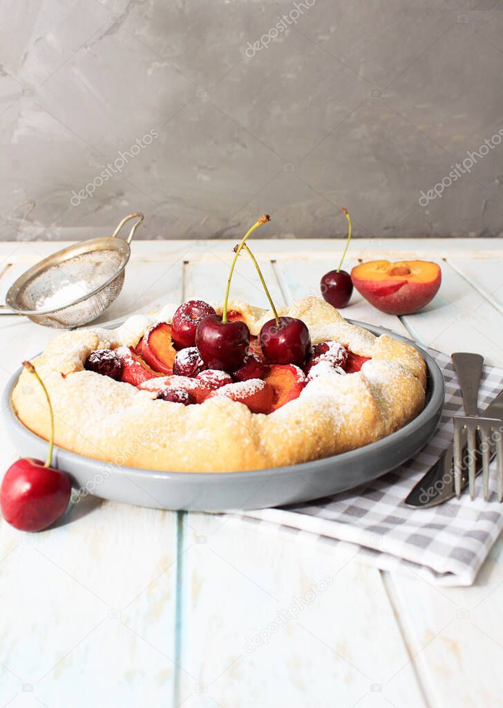 Homemade summer cake, galette with cherries and peaches with powdered sugar on a gray plate on a wooden background.