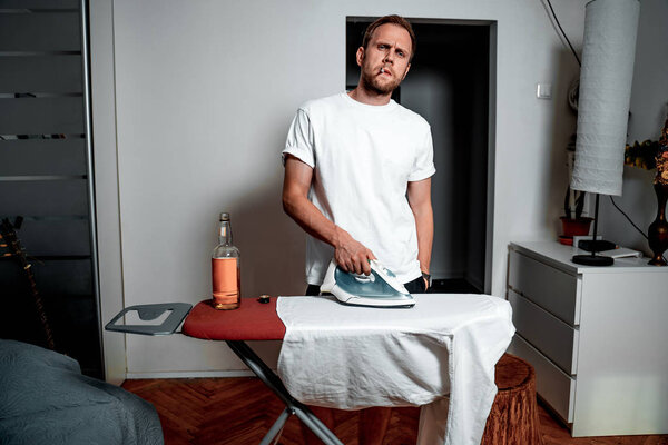 A man in a white T-shirt with a cigarette and a drink ironing his shirt