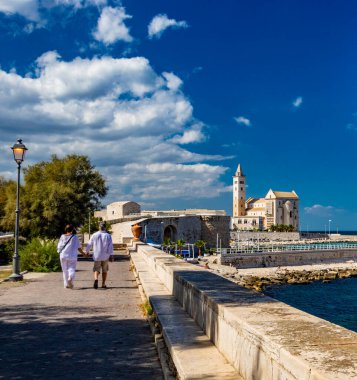İki sevgili el ele tutuşarak, Trani belediye parkında deniz manzarası ve arka planda Aziz Nicholas Pilgrim Katedral Bazilikası ile yürüyor. Puglia 'da, Bari yakınlarında, Barletta, Andria.