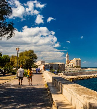 İki sevgili el ele tutuşarak, Trani belediye parkında deniz manzarası ve arka planda Aziz Nicholas Pilgrim Katedral Bazilikası ile yürüyor. Puglia 'da, Bari yakınlarında, Barletta, Andria.