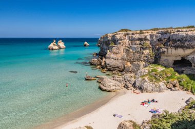 Torre dell 'Orso körfezi, yüksek uçurumları, Salento, Puglia, İtalya. Turkuaz deniz ve mavi gökyüzü. Güzel beyaz ve pembe kumdan oluşan bir kumsal. Yığınlar İki Kız Kardeş 'i çağırdı. Turistler güneşlenir..