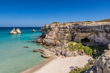 The bay of Torre dell'Orso, with its high cliffs, in Salento, Puglia, Italy. Turquoise sea and blue sky. A beach of fine white and pink sand. The stacks called the Two Sisters. Tourists sunbathe. clipart