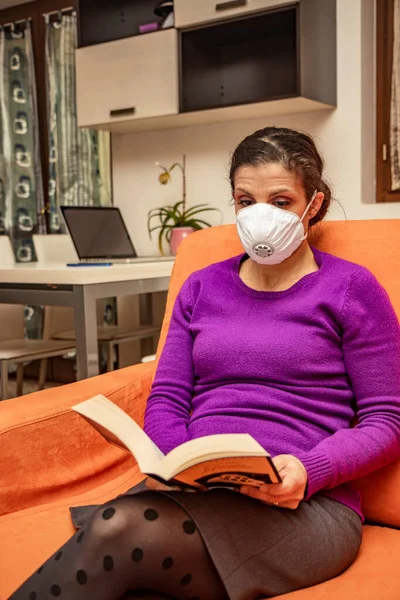 Corona Virus, Covid-19. Stay at home. Quarantine. Protection mask. A beautiful Caucasian woman forced to stay home to prevent virus infection, reads a book on the sofa to pass the time.