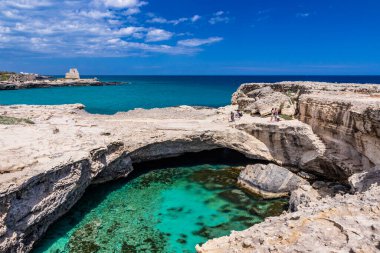 Roca Vecchia, Puglia, Salento, İtalya 'da arkeolojik alan ve turizm beldesi. Turkuaz deniz, berrak mavi gökyüzü, kayalar, güneş. Şiir Mağarası. Turistler güneşlenir. Arka plandaki gözetleme kulesi..