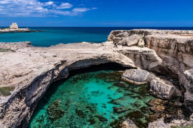 Roca Vecchia, Puglia, Salento, İtalya 'da arkeolojik alan ve turizm beldesi. Turkuaz deniz, berrak mavi gökyüzü, kayalar, güneş. Şiir Mağarası. Turistler güneşlenir. Arka plandaki gözetleme kulesi..