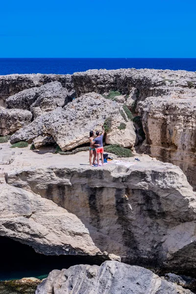 Archaeological site and tourist resort of Roca Vecchia, Puglia, Salento, Italy. Turquoise sea, clear blue sky, rocks, sun, in summer. The Cave of Poetry. A young couple takes a selfie on the rocks.