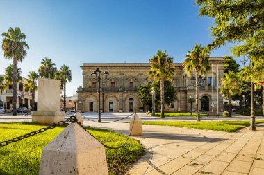 Acquarica del Capo, Presicce, Salento, Puglia, İtalya. Belediye binası ve belediye binası olan şehir meydanı. Antik taş ve tuğla bina. Çimleri ve palmiyeleri olan çiçek yatakları..