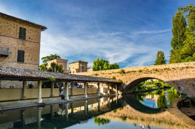1 Haziran 2019 - Bevagna, Perugia, Umbria, İtalya. Bevagna 'nın ortaçağ köyünde, nehrin üzerindeki eski yıkama ve duvarcılık köprüsü. Gün batımında mavi gökyüzü. Ağaçlar ve bitki örtüsü. Su yüzeyindeki yansıma..
