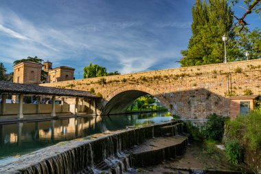 Bevagna 'nın ortaçağ köyünde, nehrin üzerindeki eski yıkama ve duvarcılık köprüsü. Perugia, Umbria, İtalya. Gün batımında mavi gökyüzü. Ağaçlar ve bitki örtüsü. Su yüzeyindeki yansıma..