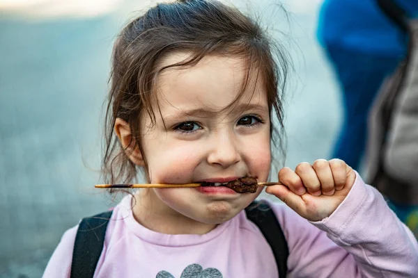 A small girl eats an Arrosticino, a skewer of mutton, a typical dish of Abruzzo cuisine. Eat with taste and appetite. To be hungry. Grilled meat, barbeque.