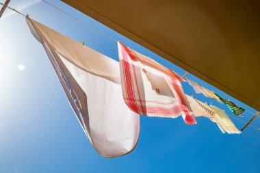 Giysiler bir evin balkonunda, mavi gökyüzünde güneşte asılı duruyor. Düşük açılı, arka ışıklandırmalı, ışıltılı ve parıltılı. Rüzgarda sallanan çarşaf, masa örtüsü ve havlu..