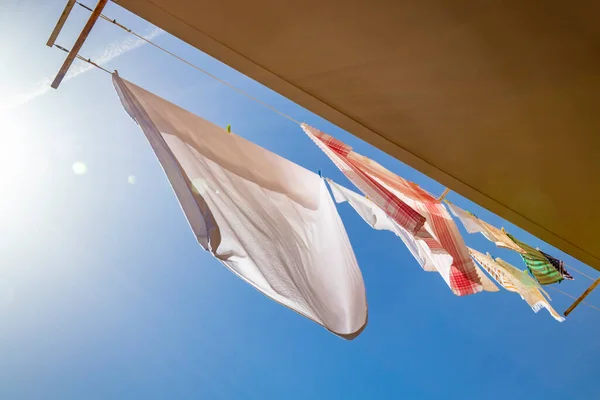Clothes hanging in the sun on the balcony of a house, in the blue sky. Low angle view, backlit, with glow and flare. Sheet, tablecloth and towel waving in the wind.