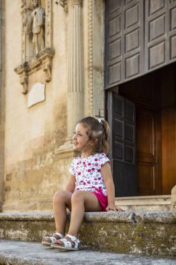 Cavallino, Lecce, Puglia, Salento, İtalya 'daki barok kilisesinin merdivenlerinde oturan güzel ve gülümseyen küçük bir kız. Fotoğrafa poz veren sevimli çocuk gülümsemeleri. Arka planda ahşap kapı.