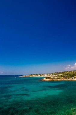 Leuca, Puglia, Salento, İtalya 'daki Apulian kıyılarının muhteşem manzarası. Turkuaz deniz, berrak mavi gökyüzü, kayalar, güneş, rüzgar, beyaz bulutlar, yazın yemyeşil bitki örtüsü. Beyaz evleri olan kalabalık bir bölge..