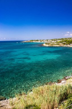 Leuca, Puglia, Salento, İtalya 'daki Apulian kıyılarının muhteşem manzarası. Turkuaz deniz, berrak mavi gökyüzü, kayalar, güneş, rüzgar, beyaz bulutlar, yazın yemyeşil bitki örtüsü. Beyaz evleri olan kalabalık bir bölge..
