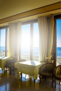 Pencereden deniz manzarası harika olan bir otelin masaları. Bir yaz günü pencereden içeri ışık girer. Tatiller, rahatlama, lüks, huzur, sükunet, dinlenme.