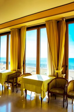 Pencereden deniz manzarası harika olan bir otelin masaları. Bir yaz günü pencereden içeri ışık girer. Tatiller, rahatlama, lüks, huzur, sükunet, dinlenme.