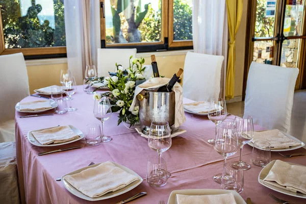 Table set for a ceremony in an Italian restaurant. Celebrations for a wedding. Basket with Champagne. Floral decorations. Pink tablecloth. Luxury, elegance, refinement.