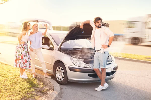 Young man in problem with broken car, while he\'s worried, his friends make selfie