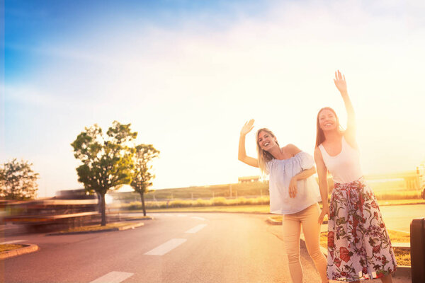 Two friends on a road trip, hipster girls hitchhiking on roadside of highway