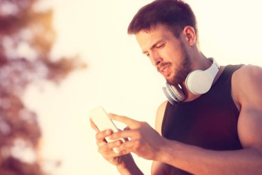 Onun iş dışarı, gülümsemek güneşli günde otururken yakışıklı genç adam akıllı telefonunu kullanarak Kulaklığımda memnun