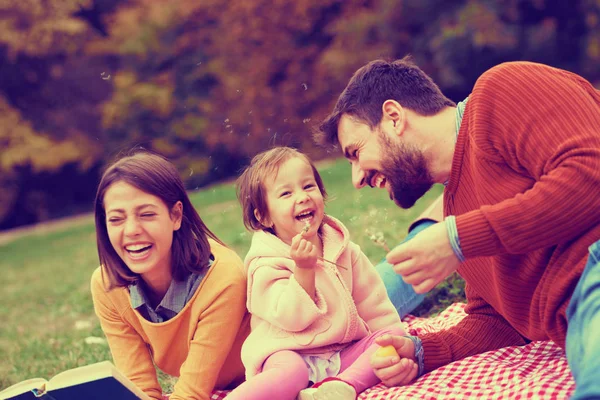 Счастливая маленькая девочка на пикнике со своими родителями, дующими одуванчиками — стоковое фото