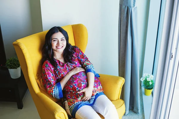 Young pregnant Indian woman sitting in armchair at home