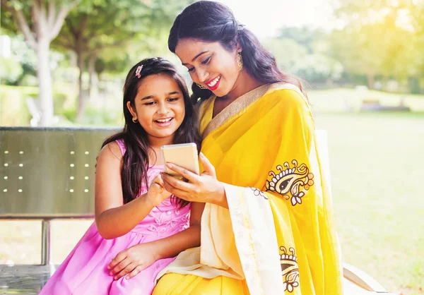 Indian mother and daughter in traditional ethnic clothes using mobile phone on a bench of the park in Delhi