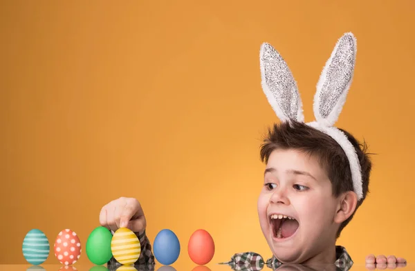 El niño con las orejas de una liebre cerca de la mesa con huevos pintados — Foto de Stock