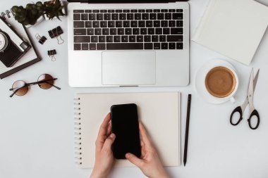 Telefon elinize. Masada bir dizüstü bilgisayar, dizüstü bilgisayar, kahve, gözlük, makas, kalem ve bir çiçek var.