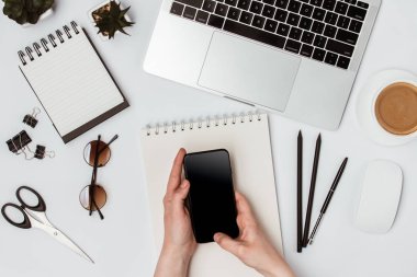 Telefon elinize. Masada bir dizüstü bilgisayar, dizüstü bilgisayar, kahve, gözlük, makas, kalem ve bir çiçek var.