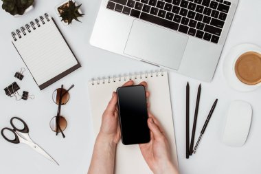 Telefon elinize. Masada bir dizüstü bilgisayar, dizüstü bilgisayar, kahve, gözlük, makas, kalem ve bir çiçek var.