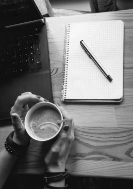 Üst Manzara. Öğrenci kadın elinde bir dilim portakalla sıcak bir fincan çay tutuyor. İş yeri, kalem ve dizüstü bilgisayarlı defter, çalışma süreci. Kahve dükkanının dikey fotoğrafı, siyah ve beyaz.