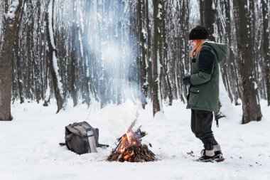 Kız kış vakti kamp ateşinin yanında duruyor. Açık havada macera tatili kavramı. Kış kampı. Karlı orman. Antika sırt çantası ve termosla çay, kahve.