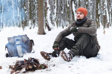 Gezgin adam kamp ateşinin yanında oturuyor. Kış zamanı. Açık havada macera tatili kavramı. Olağanüstü bir kamp. Antika sırt çantası ve termosla çay, karlı orman