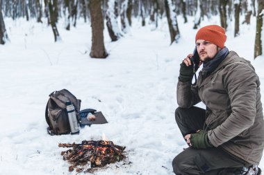 Karlı bir ormanda şöminenin yanında oturan bir adam cep telefonu kullanıyor. Cep telefonu şebekeleri. Termoslu sırt çantası. Kurtarma çağırın. Pil boşaltımı ve sinyal kaybı, turizm ve seyahatler için akıllı telefon, hayatta kalma