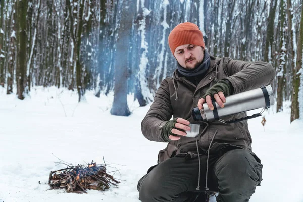 tourist man pours tea, coffee from a thermos, is sitting near camp fire in snow forest. Concept adventure active vacations outdoor. Winter camping. fleece scarf tube