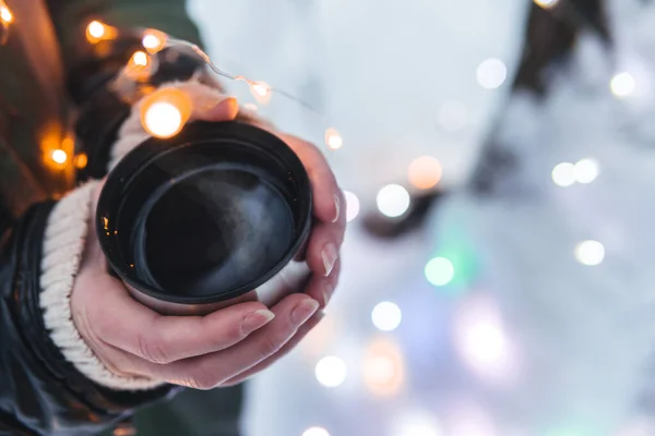 Üst manzarayı kapatın. Karlı ormanda, elinde bir fincan sıcak kahve, çay olan kız elleri. Açık havada macera tatili kavramı. Kış kampı, fotokopi alanı, renkli Noel ışıkları.