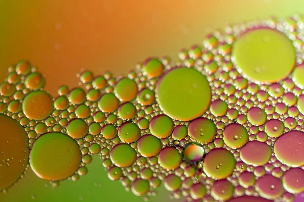 A closeup of oil droplets on a water surface with a colored background.