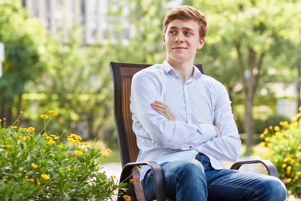 Jovem atraente homem de camisa branca sentado na cadeira marrom no parque em fundo verde — Fotografia de Stock