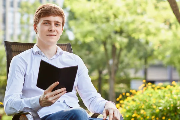 Jeune homme attrayant en chemise blanche assis sur une chaise brune dans le parc sur fond vert — Photo