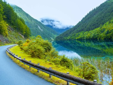 Jiuzhaigou dağları arasında, bir otoyol gölün yakınındadır. Dağlardan bir yol uzanıyor. Uzak dağlar sisle kaplıydı. Yanındaki mavi göl yeşil dağları yansıtıyor.
