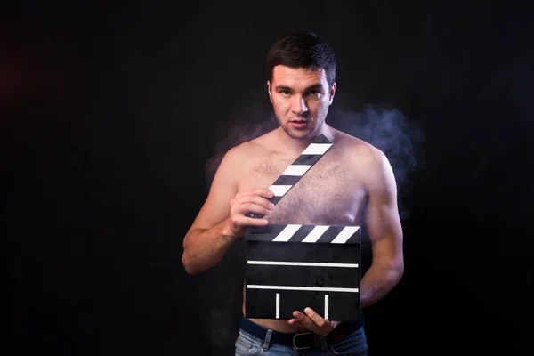 Young man of Caucasian appearance is holding a clapperboard. Portrait shot in studio on a black background with smoke and red and blue studio light.