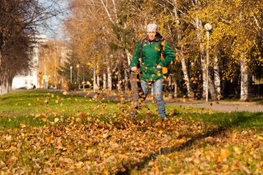 Kapıcı üniformalı bir elektrikli süpürge ile düşen sarı yapraklar yeşil çimen sokak sonbahar üzerinde bir şehir parkında kaldırır. Temiz şehir ve temiz hava, çalışma kavramı