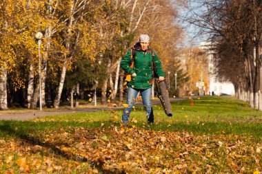 Kapıcı üniformalı bir elektrikli süpürge ile düşen sarı yapraklar yeşil çimen sokak sonbahar üzerinde bir şehir parkında kaldırır. Temiz şehir ve temiz hava, çalışma kavramı