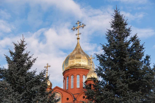 Old red brick Christian church in Kemerovo with golden and gilded domes against a blue sky and tree branches. Concept of faith in god, orthodoxy, prayer