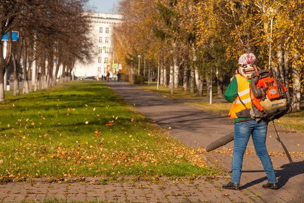 Janitor in a uniform with a vacuum cleaner removes fallen yellow leaves from green grass on an autumn street in a city park. The concept of clean city and work in the fresh air