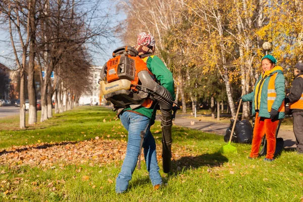 Janitors in uniform with a vacuum cleaner removes fallen yellow leaves from green grass on an autumn street in a city park. The concept of clean city and work in the fresh air